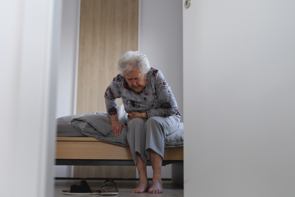 Senior woman trying to stand up from bed, feeling pain, holding abdomen.