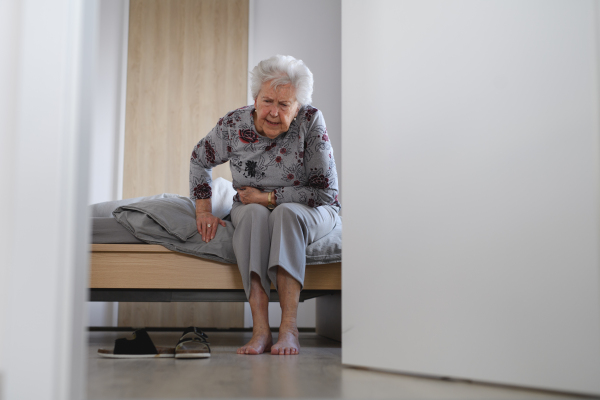 Senior woman trying to stand up from bed, feeling pain, holding abdomen.