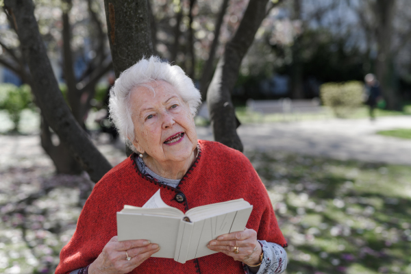 Elderly woman reading book outdoor, in park, having relaxing moment. Grandmother reading the bible in the cemetery.