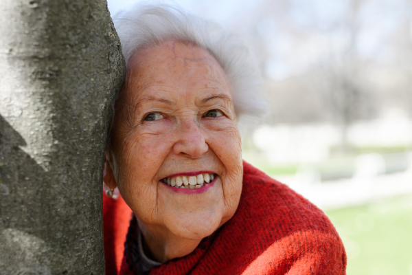 Portrait of beautiful elderly woman standing by magnolia tree, in park, having relaxing moment. Grandmother enjoying warm spring weather.