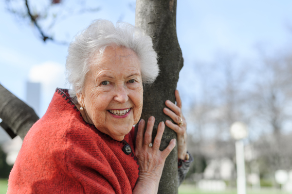 Portrait of beautiful elderly woman standing by magnolia tree, in park, having relaxing moment. Grandmother enjoying warm spring weather.