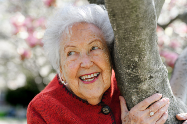 Portrait of beautiful elderly woman standing by magnolia tree, in park, having relaxing moment. Grandmother enjoying warm spring weather.