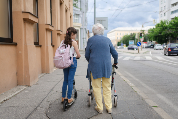 Rear view of grandma on wak with granddaughter on scooter.Granddaughter spending time with senior grandmother outdoors in city.