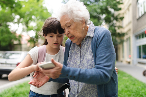 Grandma picking up young girl from school at afternoon. Granddaughter showing something on smartphone to senior grandmother, teaching her how to work with modern technology.