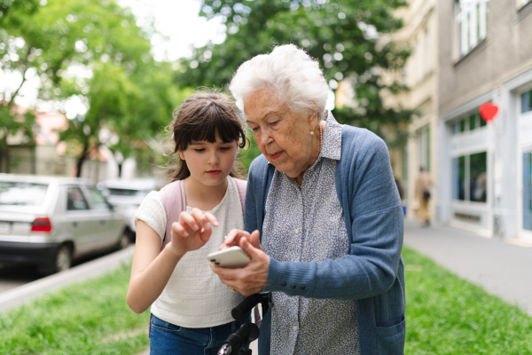 Grandma picking up young girl from school at afternoon. Granddaughter showing something on smartphone to senior grandmother, teaching her how to work with modern technology.