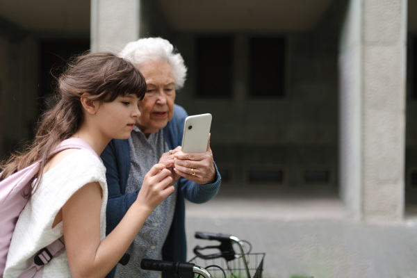 Grandma picking up young girl from school at afternoon. Granddaughter showing something on smartphone to senior grandmother, teaching her how to work with modern technology.