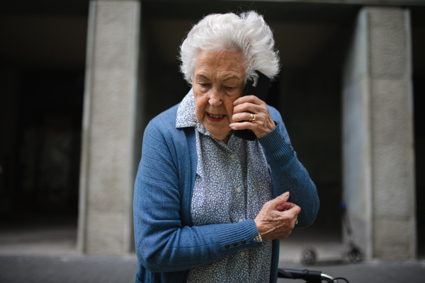 Elderly woman walking on city street with rollator while making phone call, receiving bad news.