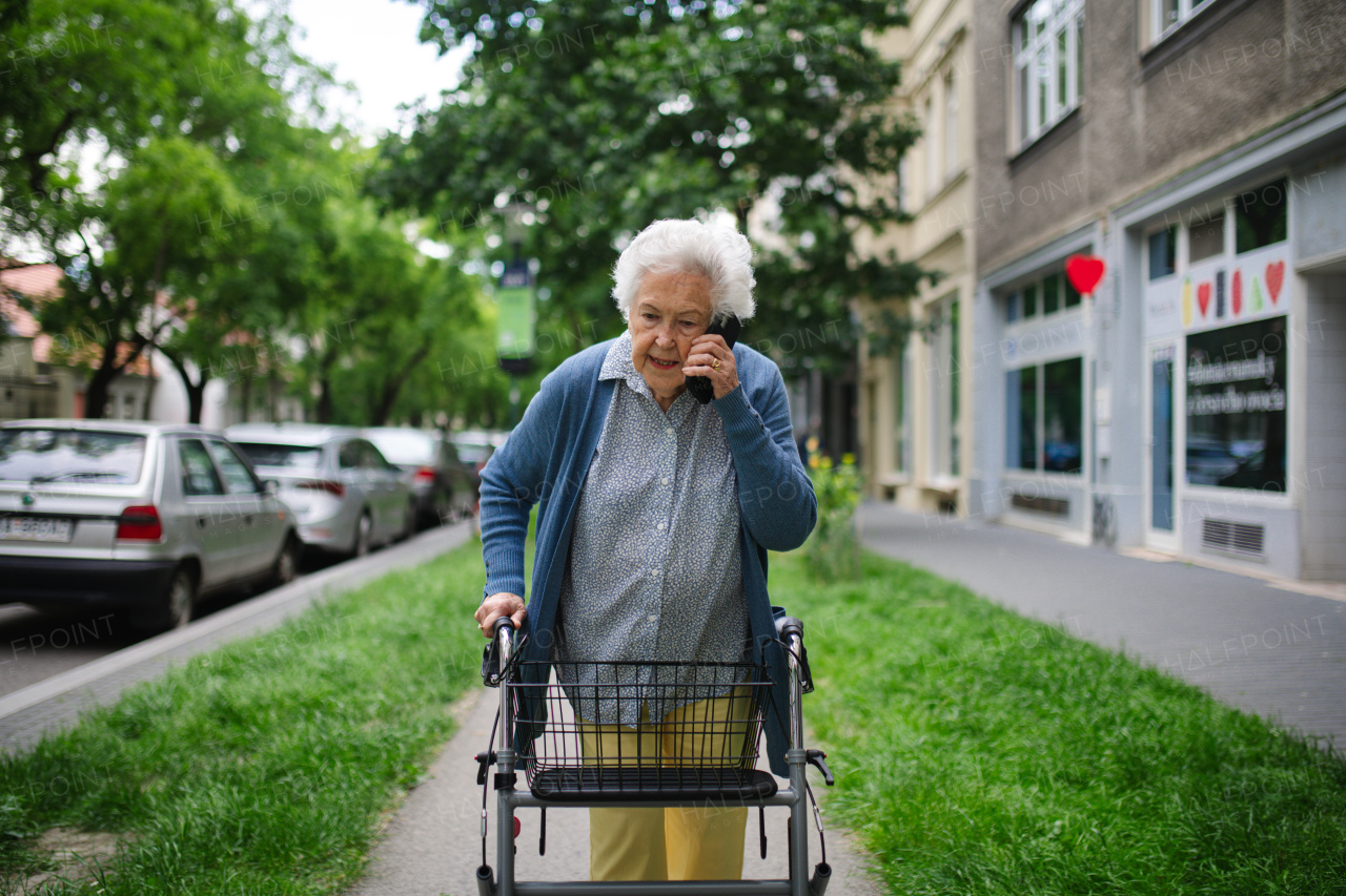 Beautiful elderly woman walking on city street with rollator, going grocery shopping to the store. Making phone call outdoors.