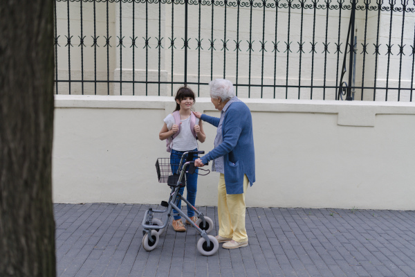 Grandma with walker picking up a young girl from school in the afternoon. Granddaughter spending time with her senior grandmother outdoors in the city after school.