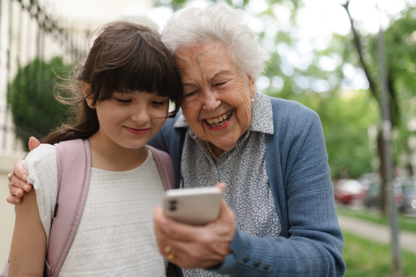 Grandma picking up young girl from school at afternoon. Granddaughter showing something on smartphone to senior grandmother.