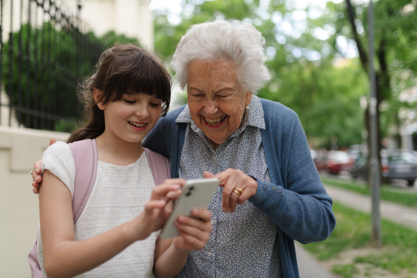 Grandma picking up young girl from school at afternoon. Granddaughter showing something on smartphone to senior grandmother.