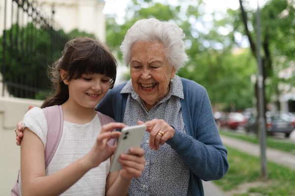 Grandma picking up young girl from school at afternoon. Granddaughter showing something on smartphone to senior grandmother, teaching her how to work with modern technology.