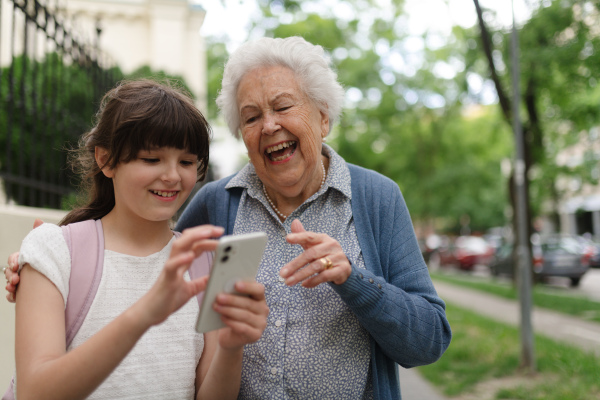 Grandma picking up young girl from school at afternoon. Granddaughter showing something on smartphone to senior grandmother.