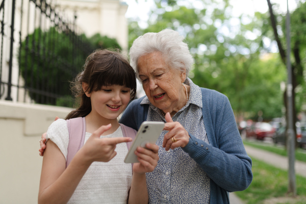 Grandma picking up young girl from school at afternoon. Granddaughter showing something on smartphone to senior grandmother.