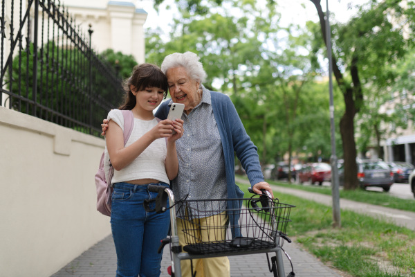 Grandma picking up young girl from school at afternoon. Granddaughter showing something on smartphone to senior grandmother, teaching her how to work with modern technology.