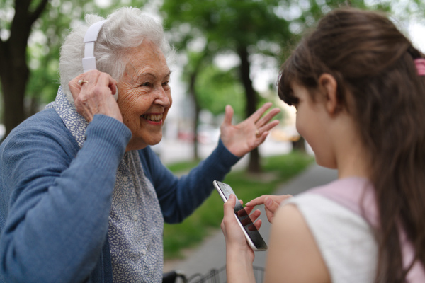 Grandma picking up young girl from school at afternoon. Granddaughter playing music on smartphone to senior grandmother with headphones.