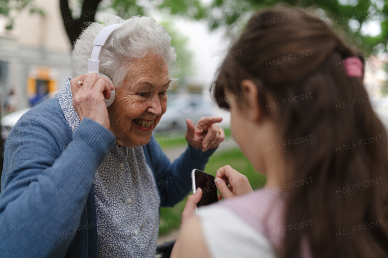 Grandma picking up young girl from school at afternoon. Granddaughter playing music on smartphone to senior grandmother with headphones.