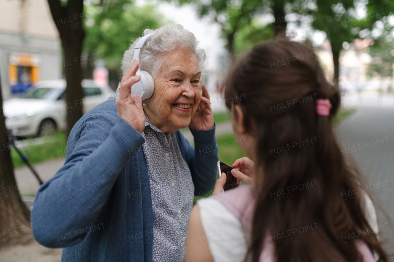 Grandma picking up young girl from school at afternoon. Granddaughter showing funny video on smartphone to senior grandmother with headphones.