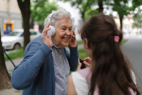 Grandma picking up young girl from school at afternoon. Granddaughter showing funny video on smartphone to senior grandmother with headphones.