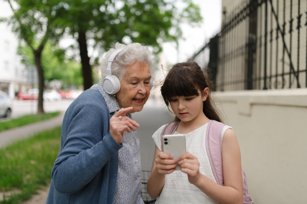 Grandma picking up young girl from school at afternoon. Granddaughter showing funny video on smartphone to senior grandmother with headphones.