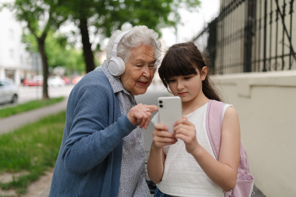 Grandma picking up young girl from school at afternoon. Granddaughter showing funny video on smartphone to senior grandmother with headphones.