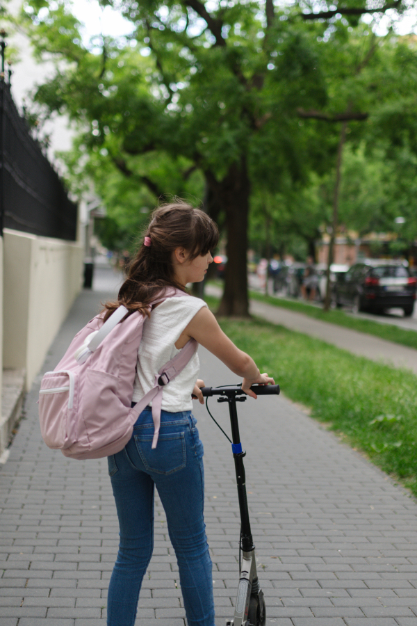 Young girl commuting on scooter to school, riding on the sidewalk with backpack on her back.