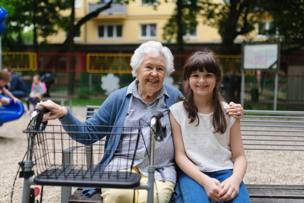 Grandma with walker picking up young girl from school at afternoon. Granddaughter spending time with senior grandmother in city park, sitting on bench in playground.