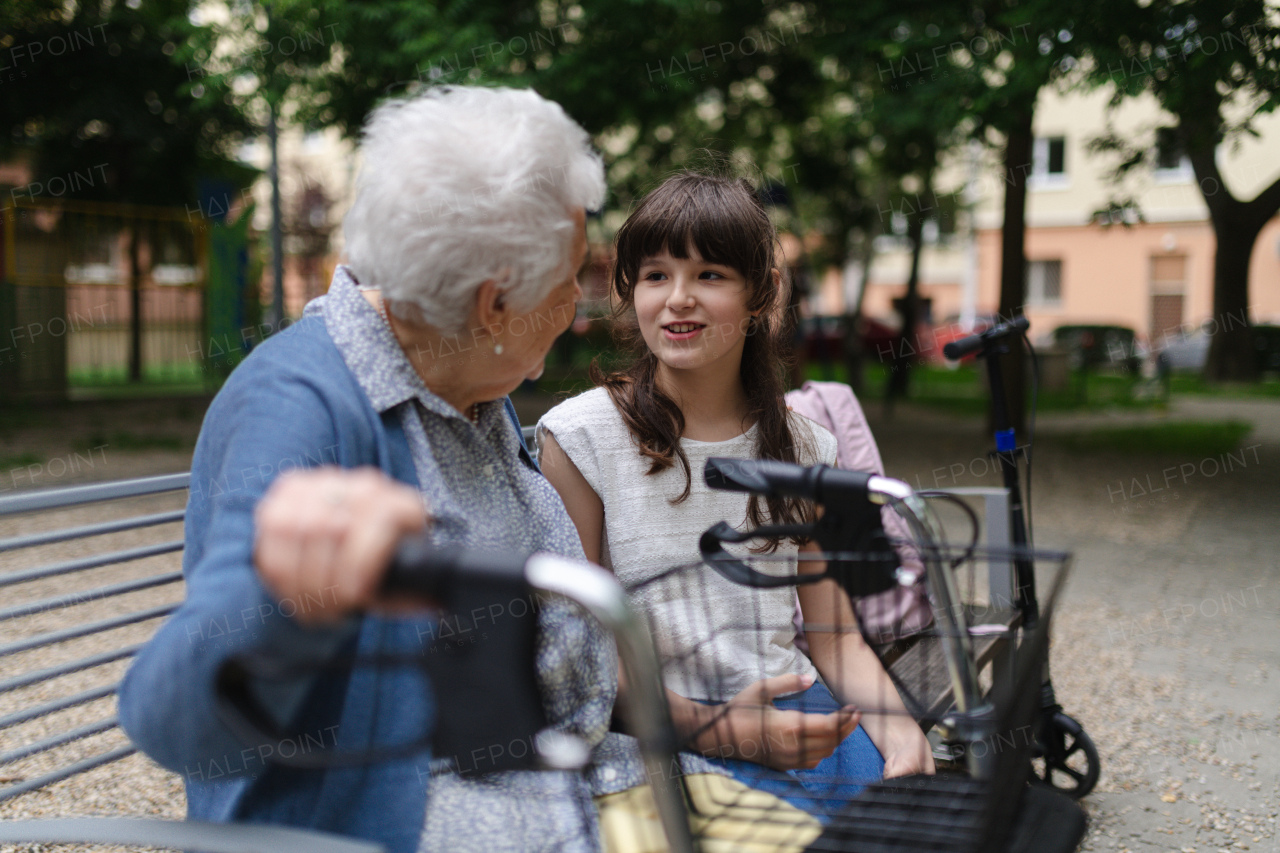 Grandma with walker spending time with granddaughter in city park, sitting on bench at children playground, talking.