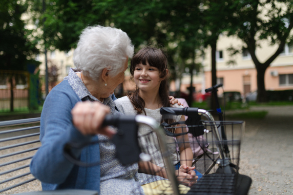 Grandma with walker spending time with granddaughter in city park, sitting on bench at children playground, talking.