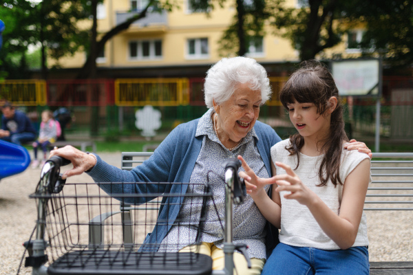 Grandma with walker picking up young girl from school at afternoon. Granddaughter spending time with senior grandmother in city park, sitting on bench in playground.