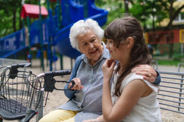 Grandma with walker spending time with granddaughter in city park, sitting on bench at children playground, talking.