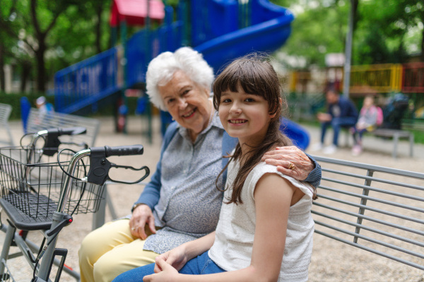 Grandma with walker picking up young girl from school at afternoon. Granddaughter spending time with senior grandmother in city park, sitting on bench in playground.