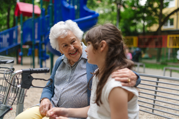 Grandma with walker spending time with granddaughter in city park, sitting on bench at children playground, talking.