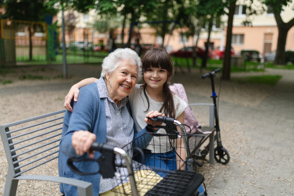 Grandma with walker picking up young girl from school at afternoon. Granddaughter spending time with senior grandmother in city park, sitting on bench in playground.