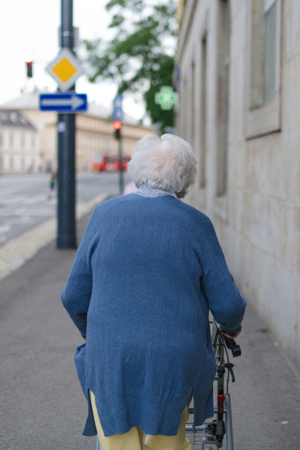 Rear view of elderly woman walking on city street with rollator, going grocery shopping to the store.