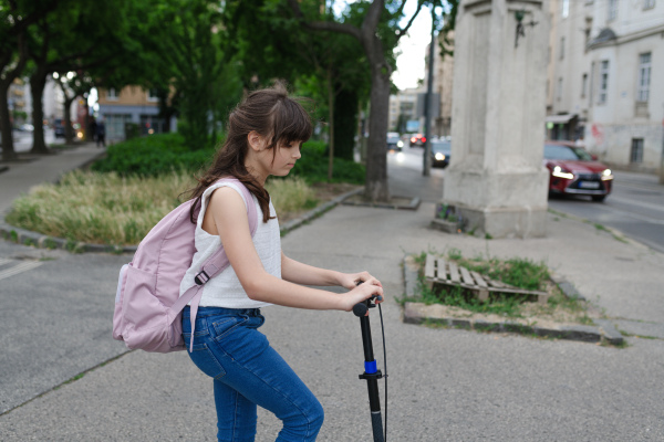 Young girl commuting on scooter to school, riding on the sidewalk with backpack on her back.