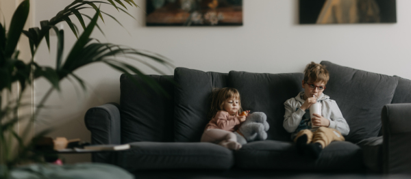 Siblings are bored during the holidays, sitting on sofa and watching television. Children spending time indoors while raining during summer break. Longer screen time during holidays. Banner with copy space