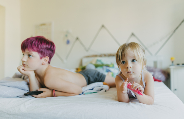 Siblings are bored during the holidays, lying on the bed and watching television. The children spending time indoors while raining during summer break. Longer screen time during holidays.