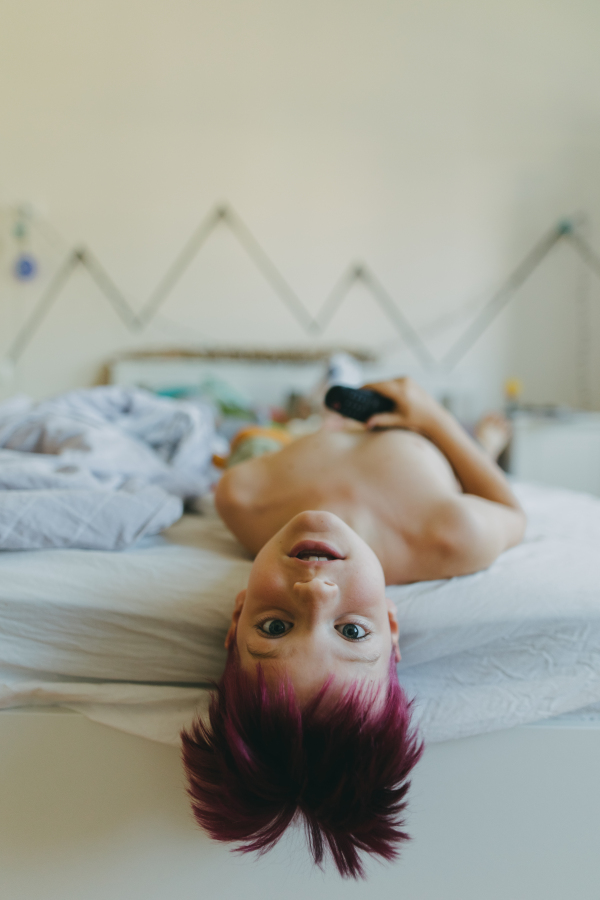 A young boy is bored at home during the holidays. Boy lying with his head hanging down from the bed, watching television. Student dyed his hair burgundy for holidays.