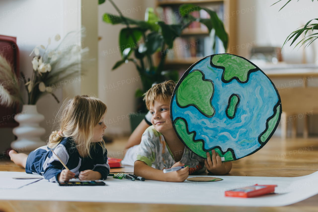 Siblings lying on stomach, painting at home with watercolors and tempera paints, creating a model of planet Earth. A creative project at home during the holidays. A young student preparing a school project on the topic of planet conservation, climate change and Earth day.