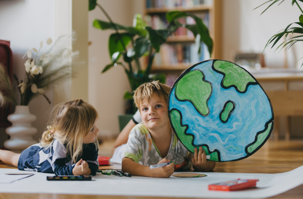 Siblings lying on stomach, painting at home with watercolors, markers and tempera paints, creating a model of planet Earth. A creative project at home during the holidays. A young student preparing a school project on the topic of planet conservation, climate change and Earth day.