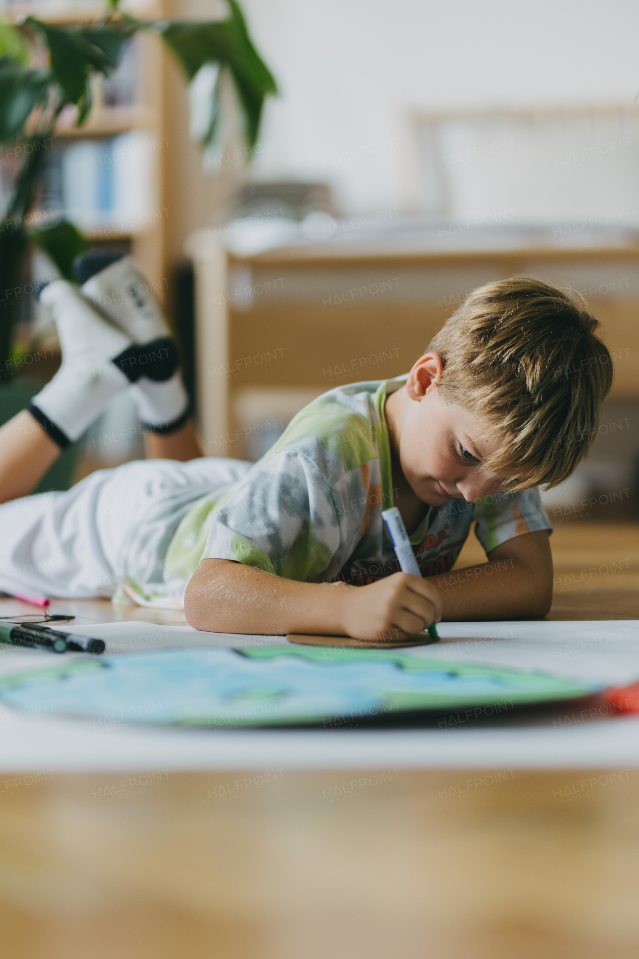 Boy lying on stomach, painting at home with watercolors and tempera paints, creating a model of planet Earth. A creative project at home during the holidays. A young student preparing a school project on the topic of planet conservation, climate change and Earth day.