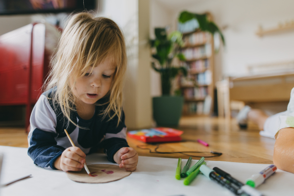 Little girl lying on stomach, painting at home with watercolors, markers and tempera paints. A creative project at home during the holidays for preschooler.