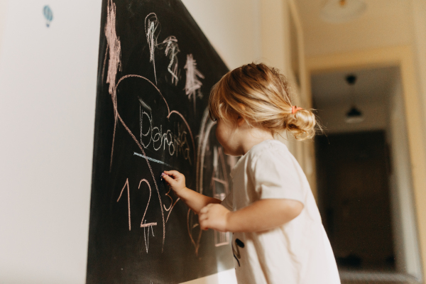 A little girl drawing on a chalkboard with chalk. The creative child is playing at home, learning to draw basic shapes and first letters.