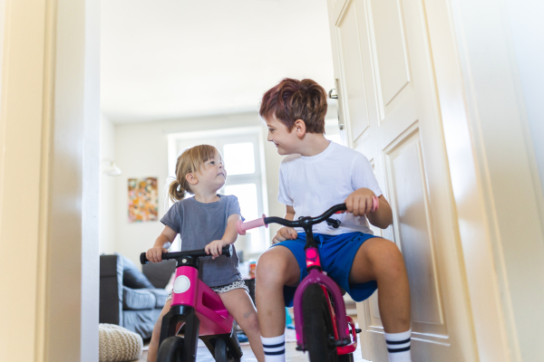 Siblings are riding bicycles indoors, in the hallway of the house. A young boy and girl are having fun during the holidays at home, racing each other on bicycles. Concept of sibling love and play.