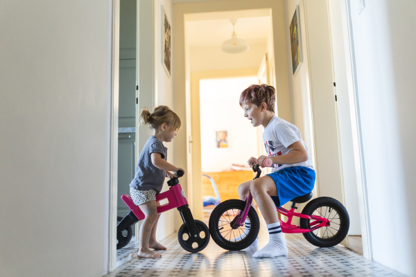 Siblings are riding bicycles indoors, in the hallway of the house. A young boy and girl are having fun during the holidays at home, racing each other on bicycles. Brother and sister sitting on bikes and facing each other. Concept of sibling love and sibling play.