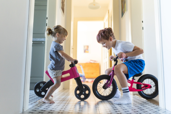 Siblings are riding bicycles indoors, in the hallway of the house. A young boy and girl are having fun during the holidays at home, racing each other on bicycles. Brother and sister sitting on bikes, facing each other. Concept of sibling love and sibling play.