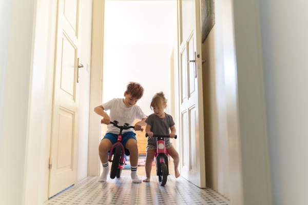 Siblings are riding bicycles indoors, in the hallway of the house. A young boy and girl are having fun during the holidays at home, racing each other on bicycles. Concept of sibling love and sibling play.