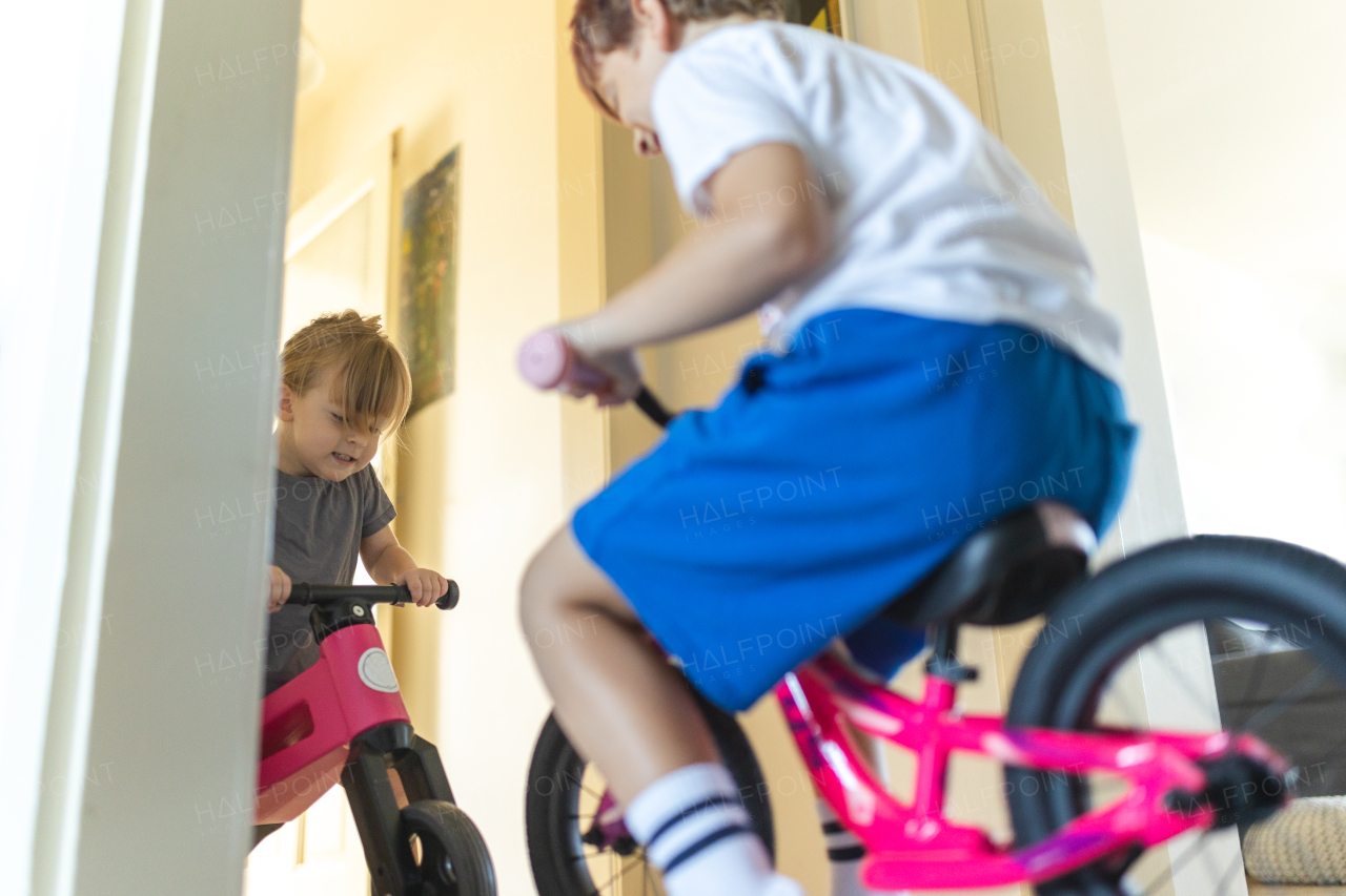 Siblings are riding bicycles indoors, in the hallway of the house. A young boy and girl are having fun during the holidays at home, racing each other on bicycles. Brother and sister sitting on bikes and facing each other. Concept of sibling love and sibling play.