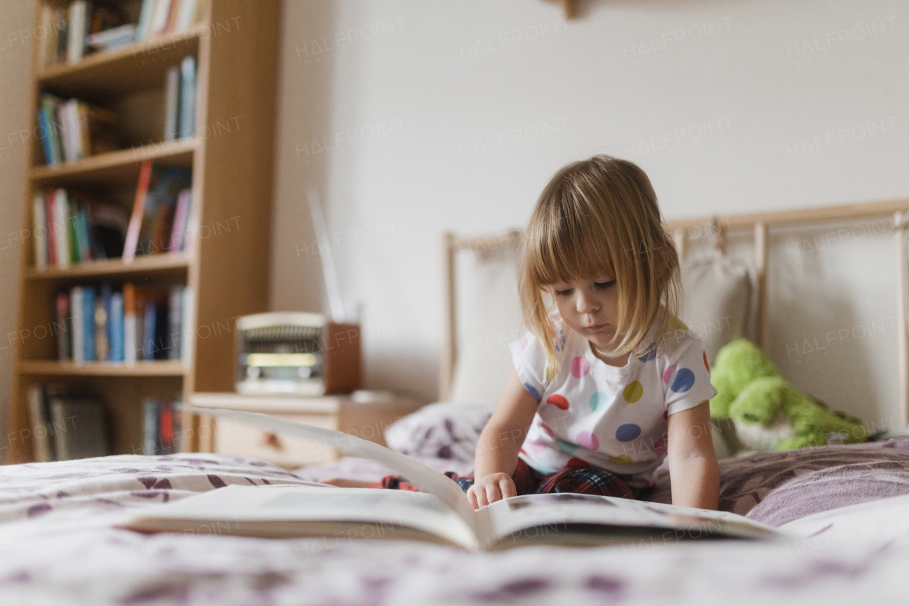 Cute little girl with bangs reading book lying on bed in her child's room.